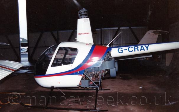 Side view of a small helicopter parked in a dark hangar, facing to the left.
The 'copter is mostly white, with a red and blue stripe running along the body and over the top of the fuselage.
The blue registration "G-CRAY" is on the white tail boom, and the red word "helicopters" is on the fairing covering the rotor mast.
The engine is partially exposed at the lower rear of the fuselage, with the whole thing sitting on black skids.
The metal doors of the hangar are slightly open, a large crack on the left of the frame revealing an arched green hangar and pale sky outside, while another couple of planes can be seen inside the dark hangar.