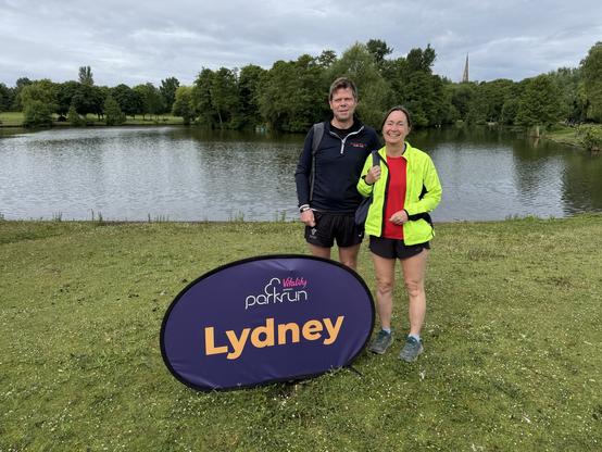 Us standing next to the Lydney Parkrun sign in front of a lake. The weather is overcast and a church spire can just be seen over the tops of the park trees in the distance.