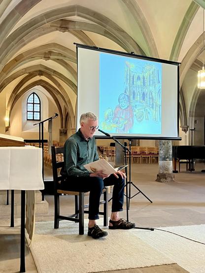 Eine Person sitzt auf einem Stuhl und liest in einer Kirche aus einem Buch vor. Hinter ihr zeigt ein großer Bildschirm eine bunte Illustration, und die Architektur zeichnet sich durch hohe Gewölbedecken aus. Im Hintergrund sind leere Stühle zu sehen.