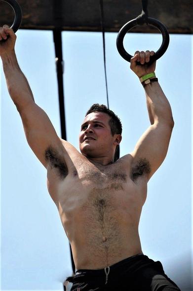A muscular white man holding on to monkey bars/rings midway through an obstacle course