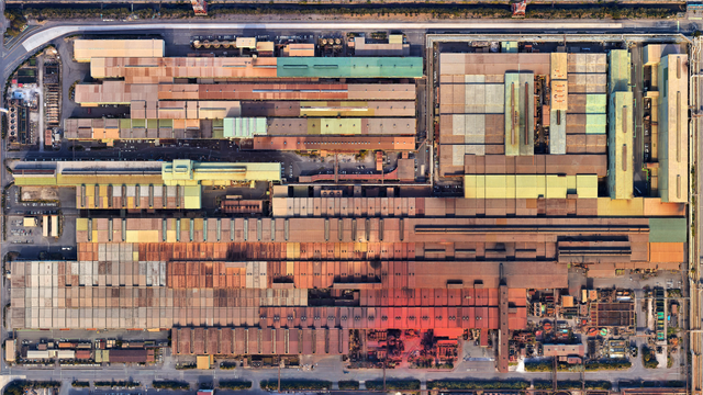 A satellite picture of a vast industrial complex: dozens of long, low factory buildings packed tightly in parallel rows, their roofs forming a patchwork of muted browns, rust reds, pale yellows, and greens. Narrow internal roads, parked vehicles, and clusters of pipes and machinery border the rectangle-shaped site.