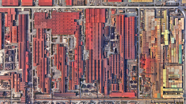 A satellite picture of a tight lattice of long, bright-red factory sheds dominates the frame, with narrow service roads threading between them; along the far right edge sits a thin strip of smaller pastel-coloured industrial roofs.
