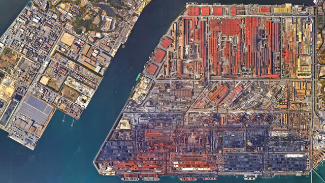 A satellite picture of a wedge-shaped industrial peninsula—top lined with red-roofed plants, centre packed with processing units, bottom covered by sprawling black ore yards and piers—separated by water from a smaller dockland zone on the left.