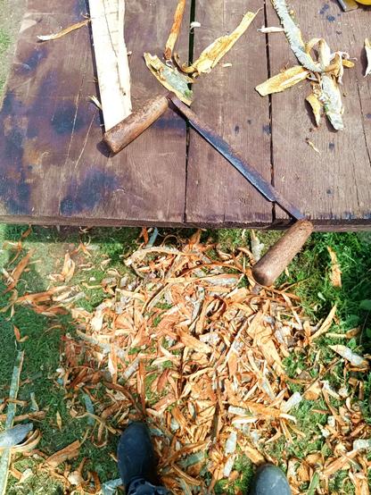 A plank table outside in the grass. A draw knife is on the table. On the ground, a pile of bark strips.