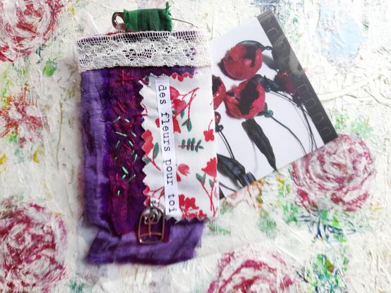A rectangular talisman on a safety pin. The collage consists of colourful silk strips in shades of purple and red, lace and intuitive embroidery in red and green. A small silver pendant shows a flower in a birdcage. ‘des fleurs pour toi’ (flowers for you) in words. It lays on a floral painting beside one of my businesscards showing a collier with red paper art flowers. Sorry for the bad photo, better ones will follow.