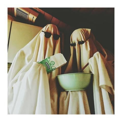 The photo is of two figures draped in white sheets (my partner and I), resembling ghosts. We are wearing sunglasses and the ghost on the left to be holding a bag of sugar and the ghost on the right is holding a bowl and a wooden spoon. You can see white kitchen cupboards in the background.