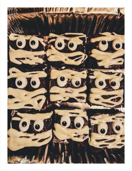 The photo is of 12 chocolate brownies decorated to look like mummies. Each brownie is topped with drizzled white chocolate to resemble bandages and two candy eyeballs. They are in a metallic baking tray.