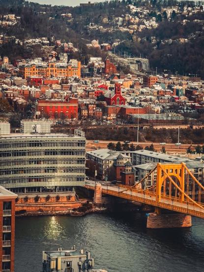 A view of the Pittsburgh north shore toward I-79N from a downtown office building featuring colorful buildings, a yellow bridge crossing a river, and a hillside with trees in the background. The scene captures a mix of architectural styles and a winter atmosphere.