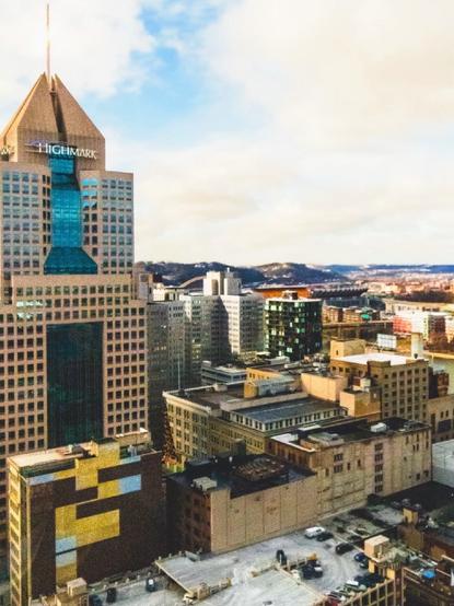 A panoramic view of the Pittsburgh skyline taken from a downtown office building featuring modern and older skyscrapers, including a prominent building marked with the "Highmark" logo. Surrounding structures exhibit a mix of architectural styles, with a backdrop of distant hills under a partly cloudy sky.