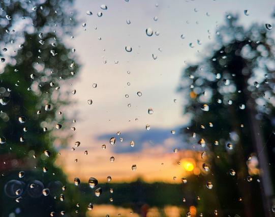 Raindrops on a window pane, in the background a blurred landscape with an orange-yellow sunset between silhouettes of trees and a lake.