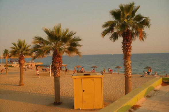 Sandy beach view with some palm trees. Between the two palm trees closest to the camera is a wooden shed kinda thing. Not sure what is inside. It’s yellow colored and goes well with the golden colors of the overall scene (golden hour). People seem to be chilling on the beach too.