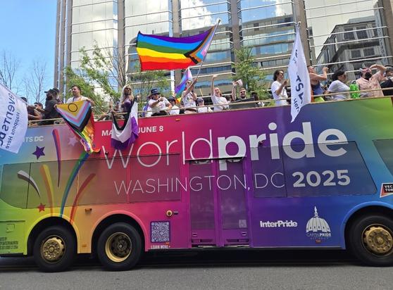 An open-top bus with "World Pride Washington, DC 2025" printed in white lettering against a gradient rainbow background, and people waving various Pride flags.