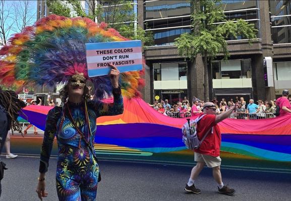 A parade marcher wearing a rainbow feather headdress with a gold sequined mask and a colorful bodysuit holds up a blue, pink, and white trans flag sign that says "These colors don't run from fascists." The extra large pride flag continues being carried in the background.