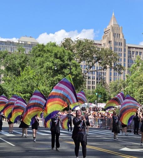 A group of color guards flag spinners march in the pride parade carrying flags with all the colors of the progress pride flag arranged in an arch.