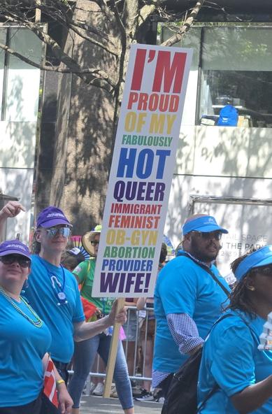 A Pride parade marcher holds a large sign in rainbow type that says "I'm proud of my fabulously hot queer immigrant feminist OB-GYN abortion-provider wife!" while pointing at the woman marching next to them.