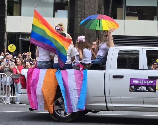Actress and singer Reneé Rapp waves to the parade crowd from the back of a white pickup truck as she holds a rainbow umbrella. Other women in the truck wave lesbian pride, trans pride, and rainbow gay pride flags.