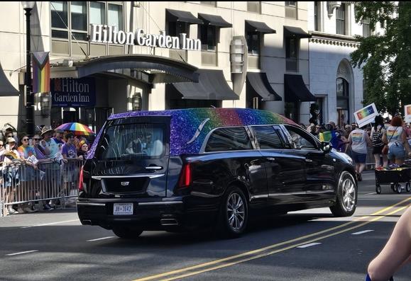 A black hearse with the top of the car decorated in gradient rainbow glitter drives in the pride parade.
