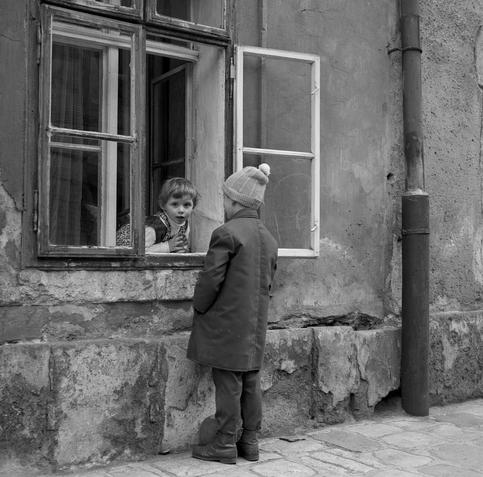 A black and white photo of a young girl looking out of a ground-floor window, directly at the photographer, who is perhaps on a 135 degree angle to her. Directly in front of her is a young boy in a coat and wooly hat.