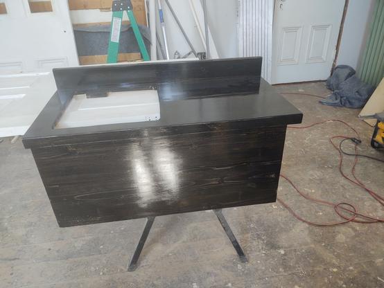 A small black wooden counter top and splash panel sitting on sawhorses. An opening for a sink has been cut into the top. Lights are reflecting off the splash