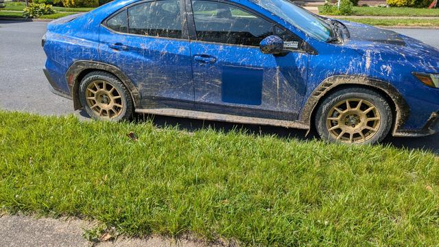 A world rally blue 2023 wrx with gold sparco wheel. The entire car is covered in clay mud.