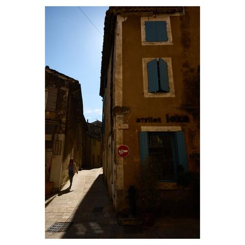 Wide angle image taken in a small village in Provence, France. The right of the frame shows a building in the shade. On the left, a small street next to it is lit by sunlight. A person is walking down the street towards the camera.