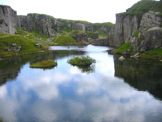 A deep and wide area of water with towering cliffs around it formed by blasting out the granite for buildings. Around the edge of the water is lots of vegetation and some on small islands as well. The sky above looks grey, although the reflection of it in the water shows fluffy clouds and blue patches in between.