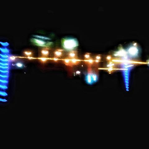 La imagen muestra una escena nocturna con una serie de luces brillantes y borrosas. En el centro, hay una línea horizontal de luces amarillas que parecen ser una fila de luces de decoración, posiblemente de una estructura o un edificio. A la izquierda, hay una columna vertical iluminada con luces azules, que podría ser parte de una estructura más grande. A la derecha, hay otra columna vertical con luces azules, similar a la de la izquierda. El fondo es predominantemente oscuro, lo que hace que las luces destaquen aún más. La imagen tiene un efecto de movimiento, lo que sugiere que la cámara estaba en movimiento o que la exposición fue larga, lo que resulta en una falta de claridad. No hay detalles claros que permitan identificar objetos o personas específicas.