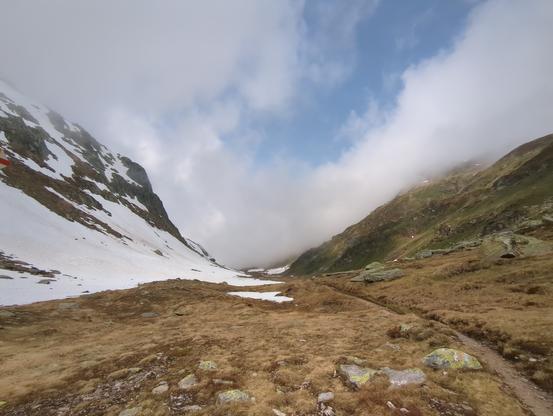 Offenes Tal mit einem breiten Talboden. Der Hang links ist noch Schneebedeckt, der rechte bereits ein wenig Grün. Rechts im Bild verläuft der Wanderweg über den braunen Talboden. In der Ferne sitzen die wolken im Tal