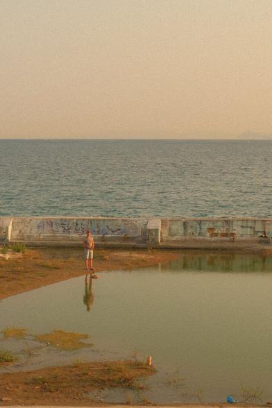 A shallow looking lake with dirt around it. A guy is standing on its edge fishing in it and his reflection is visible on the lake water. Behind him there is a cement wall, behind which is the sea! The light is kinda yellow as it’s the hour before sunset.