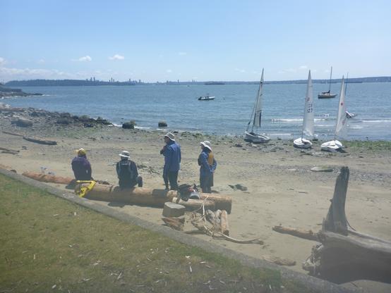 View of sailors eating lunch on the beach. Sailboats are in the background. Downtown Vancouver is seen on the opposite side of the bay.
