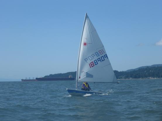 A Laser Radial sailing dinghy sailing downwind. Anchored freighters are in the background.
