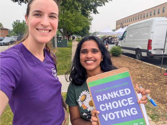 FairVote Illinois Executive Director Rebecca Williams standing alongside  a FairVote Illinois volunteer holding a clipboard that reads "Fixing politics starts with ranked choice voting" on the back.