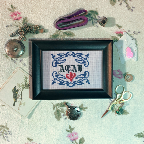a small piece of cross stitch inside a black frame surrounded by various bits of vintage looking sewing accouterments. the cross stitch is of blue art nouveau style swirling vines and leaves forming a frame around calligraphy style letters above a bright red flower. The letters read: A.C.A.B.