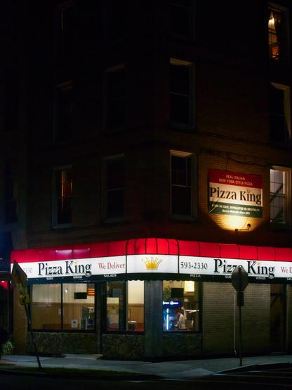 A pizzeria named Pizza King sits on a dark street corner at night. The building features a brightly lit red and white awning. Two illuminated signs are visible, displaying the restaurants name and phone number.