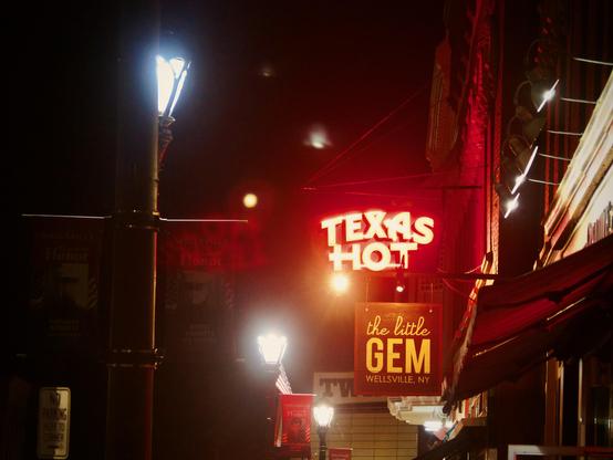 A nighttime street scene in Wellsville, NY, features glowing neon signs reading TEXAS HOT and the little GEM. Bright streetlights illuminate the area, while darkened buildings surround the vibrant signs.