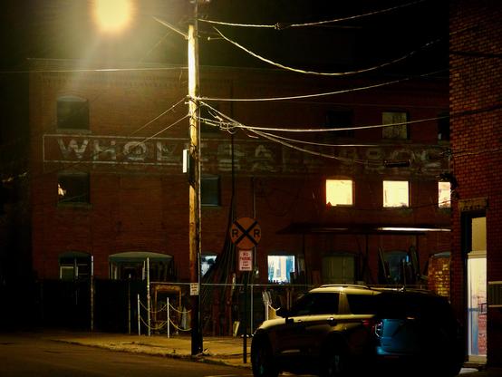 A nighttime scene on a dimly lit street. In the background, a brick warehouse with its windows illuminated casts a soft glow. Overhead, power lines crisscross the sky. In the foreground, near a parked car, is a prominent railroad crossing sign.