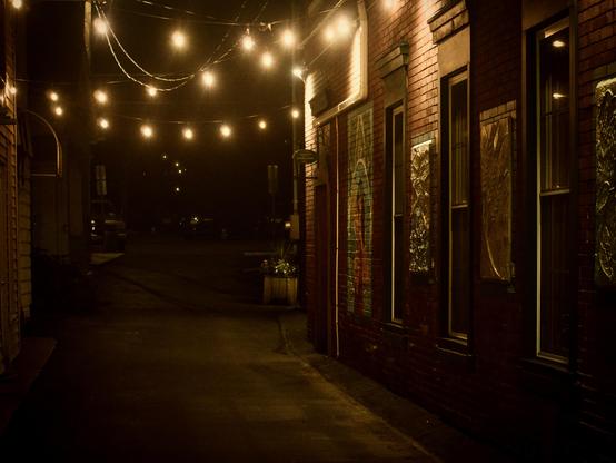 A narrow alley at night, illuminated by string lights overhead. On the right side are brick buildings with large windows and colorful mural art, both reflecting the warm glow of the lights.
