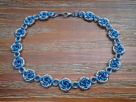 A photo of a chainmaille choker necklace on a wooden background. The necklace is a mix of blue and silver rings of various sizes. The pattern links together what chainmaillers call mobius balls, alternating between smaller and larger ones.
