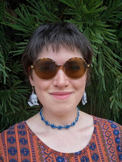 It's a selfie of me modeling my necklace, a blue and silver choker with a swirly pattern to it. I have short dark brown hair and am wearing round sunglasses and an orange and blue dress with geometric designs running down it. I'm also wearing little laser cut earrings of Shakespeare from each ear, which were also made by me!