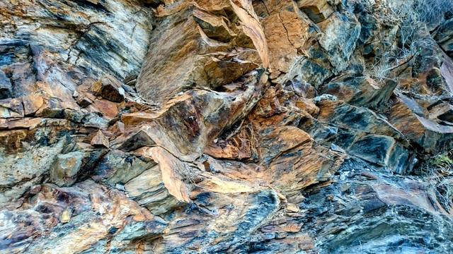 A close-up view of a rugged rock face, showing layers of grey-blue, brown, and ochre-coloured rock with sharp angles and varying textures. The rock appears weathered and fractured, with some dry, spiky vegetation clinging to the crevices on the right side. The overall impression is one of natural strength and age.