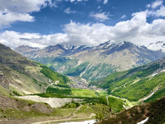 Panoramic view of a mountain valley in the Swiss Alps with lush green forests, alpine meadows, and a small village nestled in the center. Snow-capped peaks rise in the background under a partly cloudy blue sky.