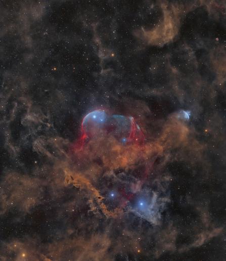 A deep-sky image showing a faint red arc near the center, representing the supernova remnant Scylla. Wisps of blue, indicating oxygen emission, mix with the red hydrogen glow across the background. In the upper right, a dim, round bluish feature marks the newly identified planetary nebula candidate, Charybdis. Numerous stars of varying brightness are scattered throughout the dark field, providing context and scale.