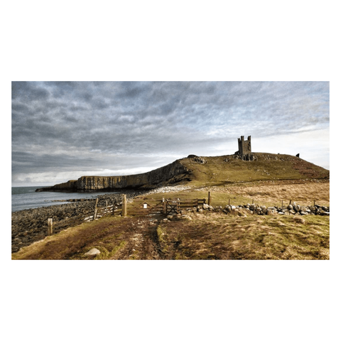 This is a landscape format colour image of the ruins of Dunstanburgh Castle on Northumberland. Set upon a promontory which juts into the North Sea, it's ruins dominate the surrounding landscape. Cliffs - home to breeding seabirds - are just visible to the left. In the foreground a rickety wooden fence and gate punctuate the image, and mirror the shape of the promontory and castle in the background. The sky is moody, and the overall palette is of muted greens, greys, and blues.