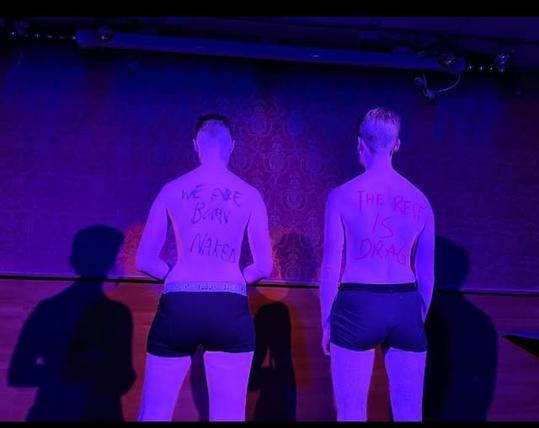Two people standing on stage with their back to the audience. Something is written on their backs.