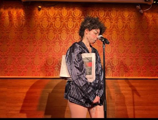 Person in pyjamas with a newspaper under their arm in front of a mic on stage.