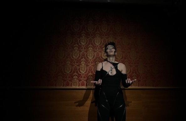 Person on stage in long black gown holding the arms open to the side of the body while their eyes are closed.