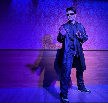 Person with outfit from the movie Matrix on stage. Holding both hands up as if to clam someone down.