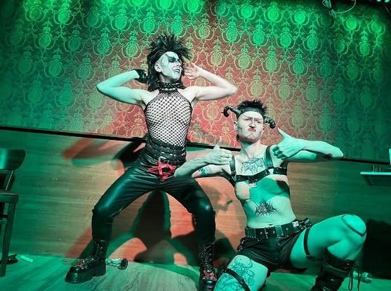 Two people on stage. One with black leather pants and a black wig twerking. The other person is wearing devil's horn is kneeling showing the audience two thumbs up.