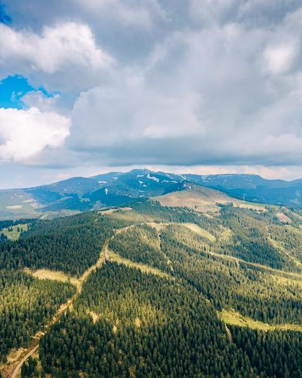 Auf dem Bild ist eine Luftaufnahme einer bergigen Landschaft zu sehen. Im Vordergrund erstreckt sich ein dicht bewaldetes Gebiet mit überwiegend Nadelbäumen. Zwischen den Bäumen verlaufen mehrere schmale, geschwungene Wege, die sich durch den Wald ziehen.
Im Mittelgrund steigt das Gelände sanft an und wird lichter, mit weniger Bäumen und mehr offenen, grünen Flächen. Ganz im Hintergrund erhebt sich ein Gebirgszug mit dunklen Berghängen, auf denen vereinzelt noch Schneefelder zu sehen sind. Die Berggipfel sind größtenteils abgerundet, nicht schroff, und teilweise mit Wäldern bedeckt.
Darüber spannt sich ein dramatischer Himmel mit großen, teils dunklen Wolken, durch die aber an einigen Stellen blauer Himmel hindurchscheint – was dem Bild eine leicht mystische, aber dennoch friedliche Stimmung verleiht.