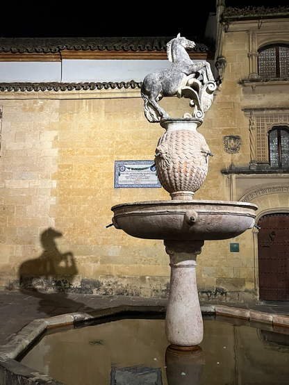 Photo of the fountain at Córdoba's Plaza del Proto at night; the horse figure on top is casting a shadow on the wall behind.